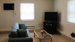 A living room with a sofa, a coffee table, a television on a stand, and a window with blinds. - Shore Bird - 1 BR Studio Cottage with open floor plan. NO LINENS PROVIDED (Chincoteague Island)