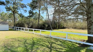 A fenced yard with a grassy area, trees, and a building partially visible. - Shore Bird - 1 BR Studio Cottage with open floor plan. NO LINENS PROVIDED (Chincoteague Island)