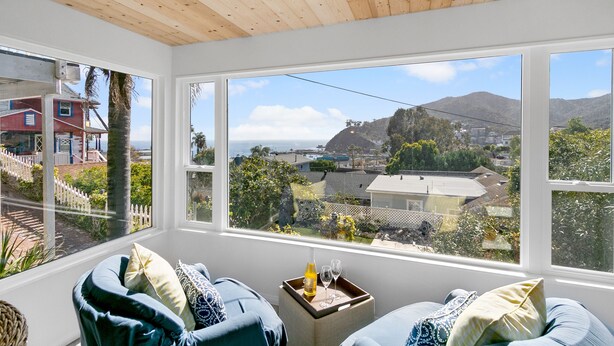 A room with a wooden ceiling, large windows offering a view of a coastal landscape, and a seating area with a tray table and two chairs. - 124H Unit 2: Cozy 2 Bedroom Unit, Stunning Views, BBQ, GOLF CART (Avalon)