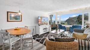 A modern living room with a dining area, large windows offering a scenic view, and a television on a white cabinet. - 124H Unit 2: Cozy 2 Bedroom Unit, Stunning Views, BBQ, GOLF CART (Avalon)