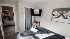 A hotel room with a bed, a TV on a stand, a painting on the wall, and a bathroom visible through an open door. - 6BR Downtown Duplex Walk to Gore & Main St | Porches, Game Room | Sleeps 12 (North Creek)