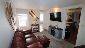 A living room with a brown leather sofa, a TV mounted on the wall, and a kitchen visible through an open doorway. - 6BR Downtown Duplex Walk to Gore & Main St | Porches, Game Room | Sleeps 12 (North Creek)