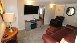 A living room with a flat-screen TV, a fireplace, a leather sofa, and a lamp. - 6BR Downtown Duplex Walk to Gore & Main St | Porches, Game Room | Sleeps 12 (North Creek)