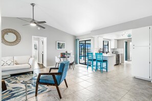 A spacious living area with a sofa, a coffee table, and a blue chair. There is a dining area with a table and chairs, and a kitchen with white cabinets and a window.