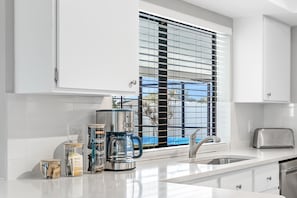 A modern kitchen with white cabinetry, a stainless steel coffee maker, a toaster, and a window with blinds.