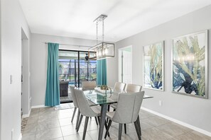 A modern dining area with a glass table, grey chairs, teal curtains, and artwork on the walls.