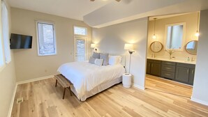 A bedroom with a bed, a bench, a ceiling fan, a TV, and a bathroom with a sink and mirror. - Enjoy Luxury & Games With A View (Maggie Valley)