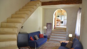 A living room with a staircase, a blue sofa, a wooden coffee table, and a kitchen visible through an archway. - Mas de Saudon (Soyans Auvergne-Rhône-Alpes)