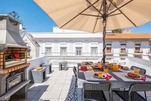 An outdoor dining area with a table set for breakfast, a grill, and a view of a white building with balconies. - Townhouse with BBQ rooftop in Tavira (Tavira)