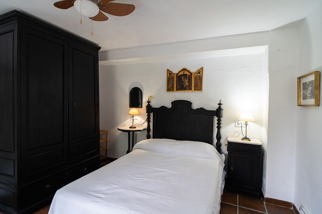 Une chambre avec une grande armoire en bois foncé, un lit avec une literie blanche, deux tables de chevet avec des lampes et un ventilateur de plafond.