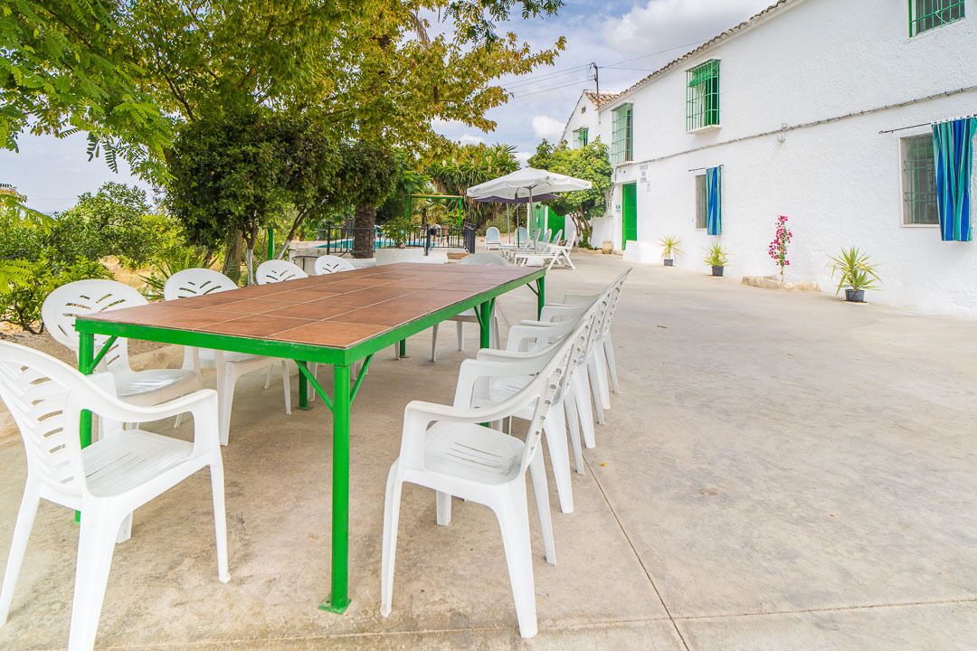 Un patio al aire libre con una larga mesa de madera y sillas blancas de plástico, rodeado de follaje y un edificio blanco con contraventanas verdes.