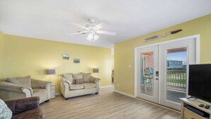 A living room with two beige sofas, a ceiling fan, a TV on a wooden stand, and a sliding glass door leading to a balcony. - Coral Breeze (Fort Morgan)