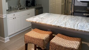Une cuisine avec des meubles blancs, un plan de travail en marbre, deux tabourets en osier et des luminaires suspendus.