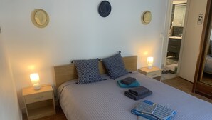 Una cama bien hecha con colcha gris, dos almohadas estampadas y toallas dobladas. Hay lámparas de noche sobre mesitas de madera.