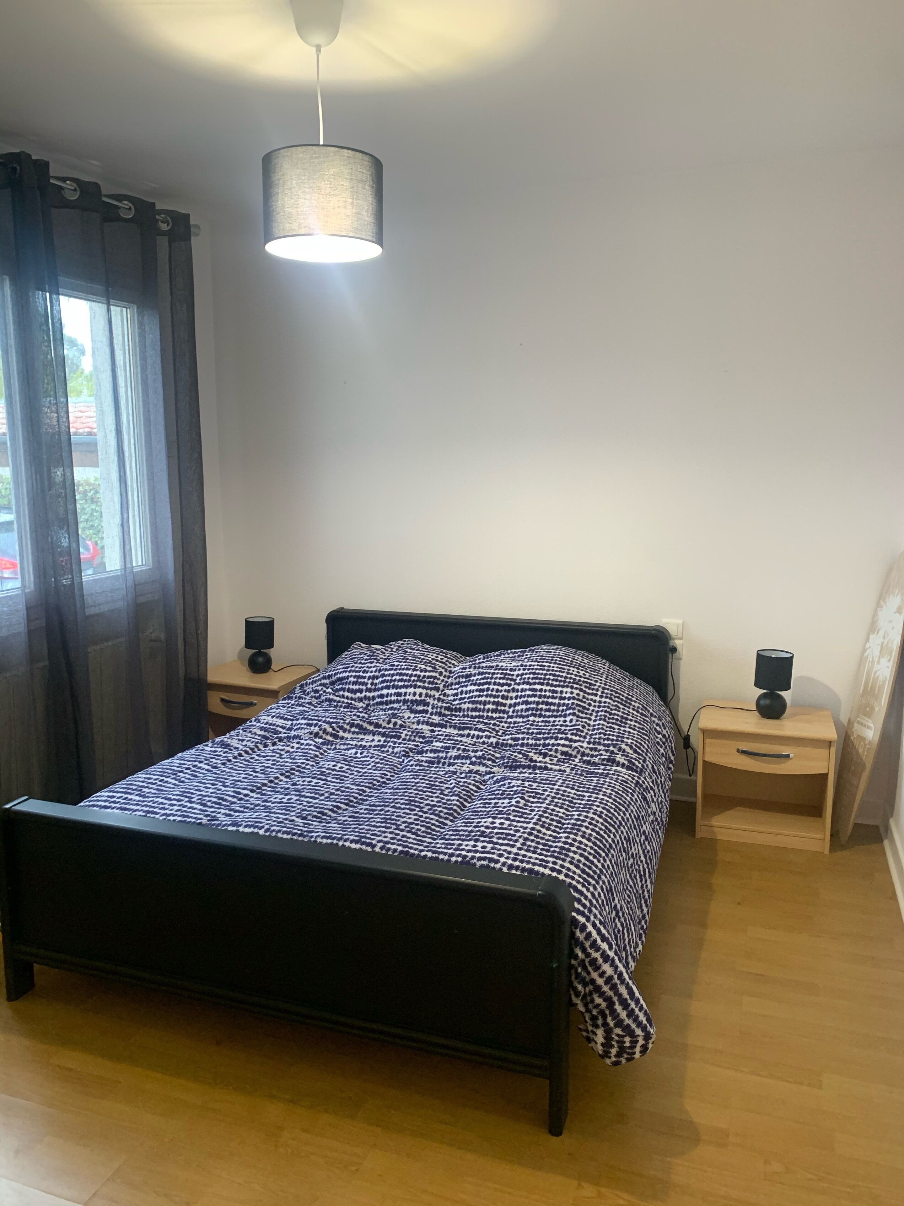 Une chambre à coucher avec un lit, deux tables de chevet avec des lampes, un luminaire suspendu et une fenêtre avec des rideaux.