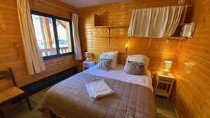 A wooden cabin bedroom with a bed, bedside lamps, a window with curtains, and a bench.