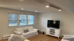 Un salon moderne avec un canapé blanc, un téléviseur posé sur un meuble blanc et une fenêtre équipée de stores.