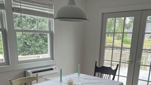 Un coin repas avec une table dressée pour quatre personnes, entouré de chaises blanches, d’un luminaire blanc au plafond et de grandes fenêtres offrant une vue sur la verdure à l’extérieur.