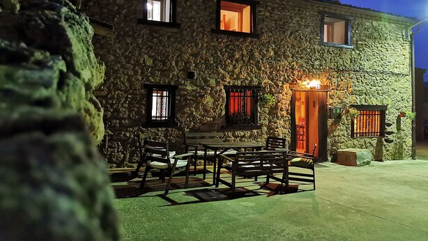 A stone building with a tiled roof, a wooden door, and a small outdoor seating area with a table and chairs. - Casa Rural de Alquiler Completo (Ligos)