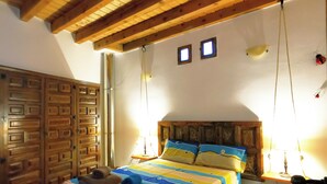 A bedroom with a wooden ceiling, a bed with a striped bedspread, a wooden wardrobe, and two small windows. - Casa Rural de Alquiler Completo (Ligos)