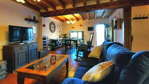 A cozy living room with a television, a coffee table with decorative items, a sofa with cushions, and a dining area with chairs and a table. - Casa Rural de Alquiler Completo (Ligos)