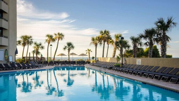 A swimming pool with lounge chairs and palm trees, surrounded by a building. - Serenity by the Sea. Oceanfront. (Myrtle Beach)