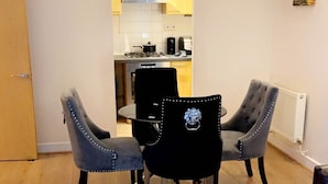 A dining area with a table and four chairs, a remote control on a table, and a kitchen in the background. - By the seaside (London)