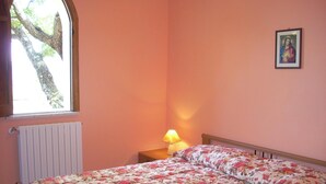 Une chambre à coucher avec une couvre-lit à motifs floraux, une table de chevet avec une lampe, un tableau encadré au mur et une fenêtre donnant sur un arbre.