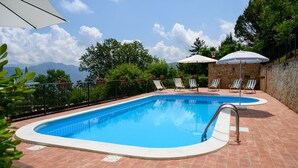 Une piscine rectangulaire, bordée d’une terrasse carrelée, entourée d’un mur de pierre et d’une clôture en métal. On y trouve deux parasols blancs et plusieurs chaises longues.
