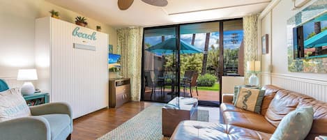 A living room with a sofa, a chair, a glass door leading to a patio, a ceiling fan, and a sign on the wall.