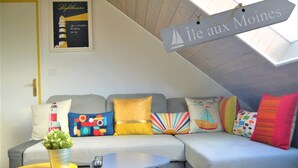 Ein gemütliches Wohnzimmer mit einem grauen Sofa, bunten Kissen, einem kleinen gelben Tisch und einem Schild mit der Aufschrift "Île aux Moines".