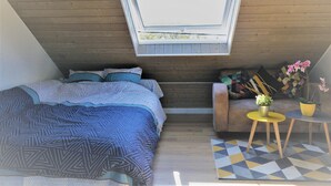 Ein kleiner, gemütlicher Raum mit schrägen Wänden, einem Bett mit gemusterten Textilien, einer Couch mit Kissen, einem Tisch mit einer Pflanze und einem Teppich mit geometrischem Muster.