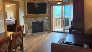 Una sala de estar con chimenea de piedra, un televisor de pantalla plana montado encima, un comedor con mesa y sillas, y un balcón con vistas al exterior.