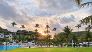 A grassy area with a pool, palm trees, and a building with multiple windows in the background. - Beach Class Muro Alto - Bangalô com Vista Para Piscina e mar (Ipojuca)