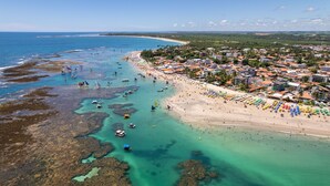An aerial view of a coastal town with a beach, clear waters, and boats. - Beach Class Muro Alto - Bangalô com Vista Para Piscina e mar (Ipojuca)