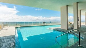 En swimmingpool med utsikt över havet, omgiven av ett staket och ett takförsett område med bord och stolar.