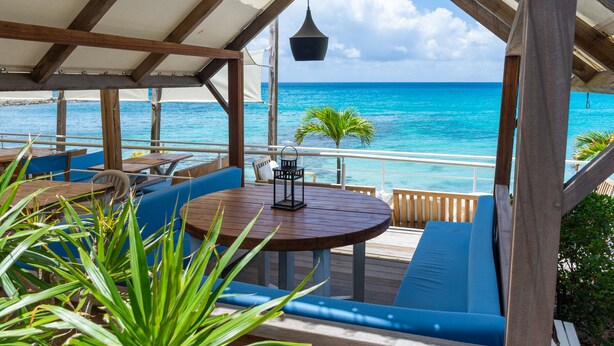 An outdoor seating area with wooden tables and blue cushioned chairs, overlooking a turquoise sea. - Villa Ocean - Indulge Yourself in Paradise (Indigo Bay)