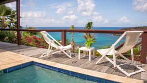 A wooden deck with two white lounge chairs and a small table, overlooking a pool and a scenic ocean view. - Villa Ocean - Indulge Yourself in Paradise (Indigo Bay)
