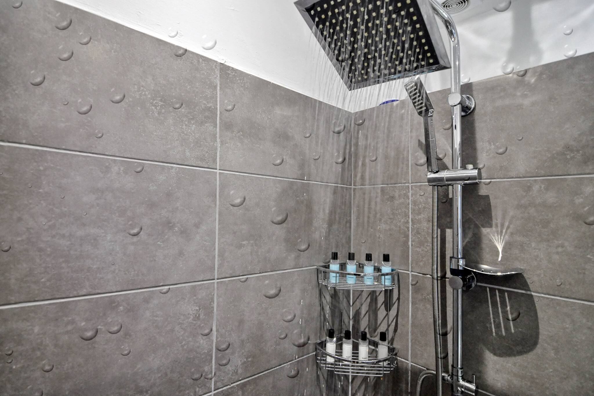 Une douche moderne dotée d’un pommeau de douche à pluie, d’un pommeau de douche à main et d’une étagère pour les articles de toilette.