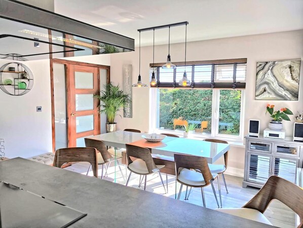 A modern dining area with a large table, chairs, and pendant lights. There is a window with a view of greenery outside.
