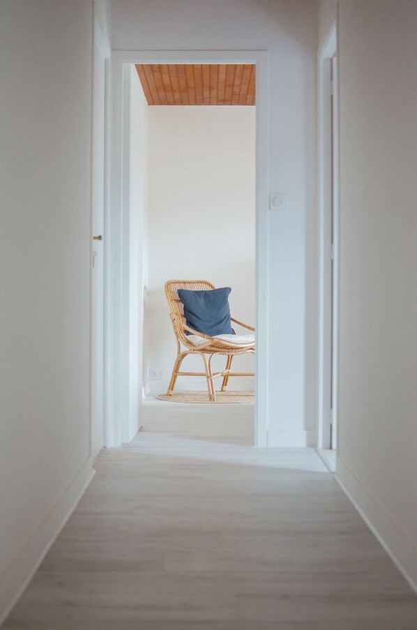 A narrow hallway with a wooden floor leading to a room with a wicker chair and a blue pillow. - Just a stone's throw from the beaches, a house with a large fenced-in yard (Cléder)