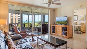 Un salon comprenant un canapé, un fauteuil et une table basse. On y trouve également un téléviseur posé sur un meuble en bois. La pièce donne sur un balcon d’où l’on peut admirer l’océan.
