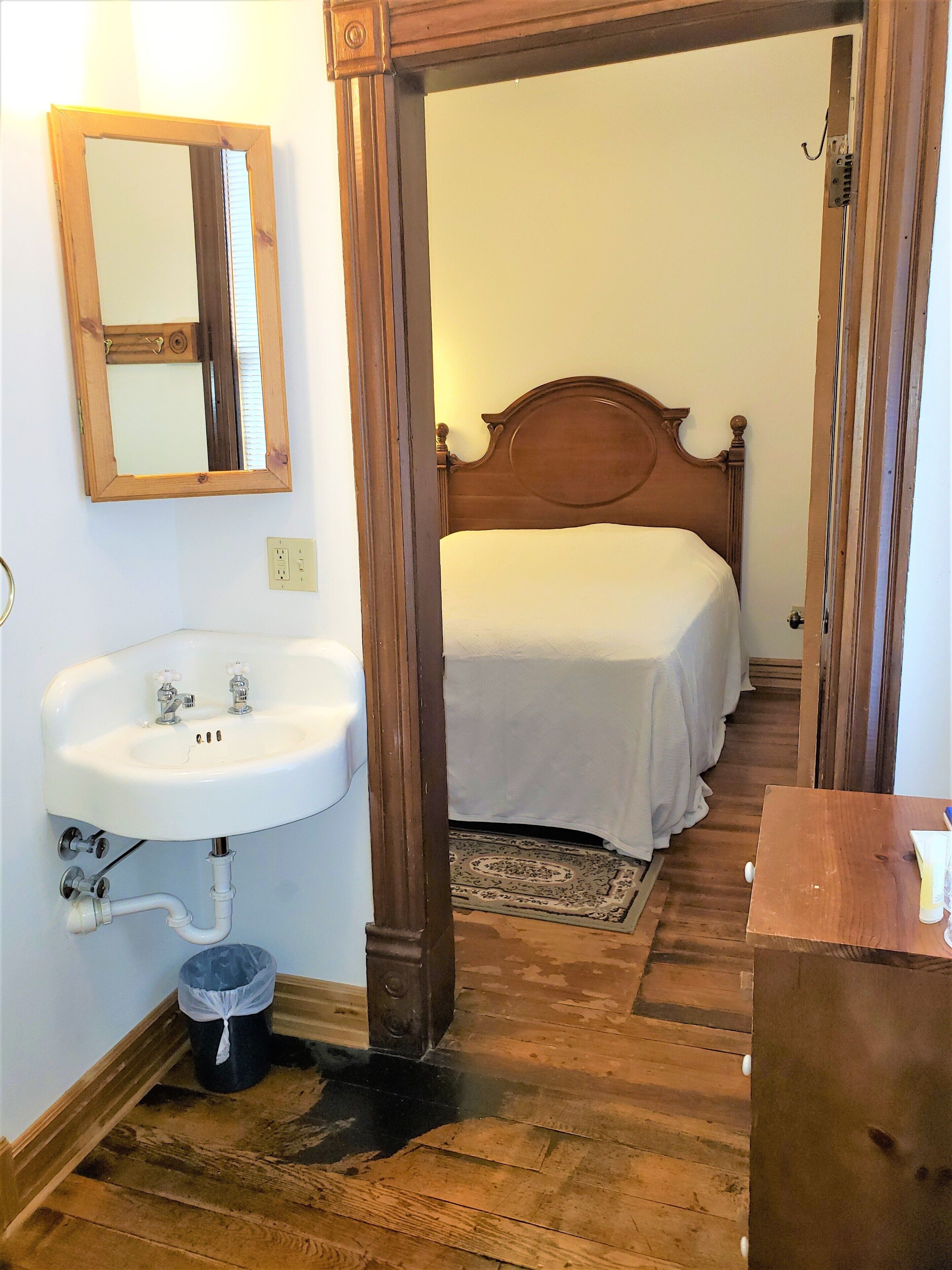 Una camera da letto con testiera in legno, copripiumino bianco, pavimento in legno, lavabo bianco con due rubinetti e un piccolo cestino.