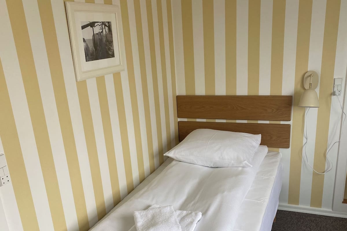 A single bed with white linens and pillows, a wooden headboard, a framed picture on the wall, and a bedside lamp.