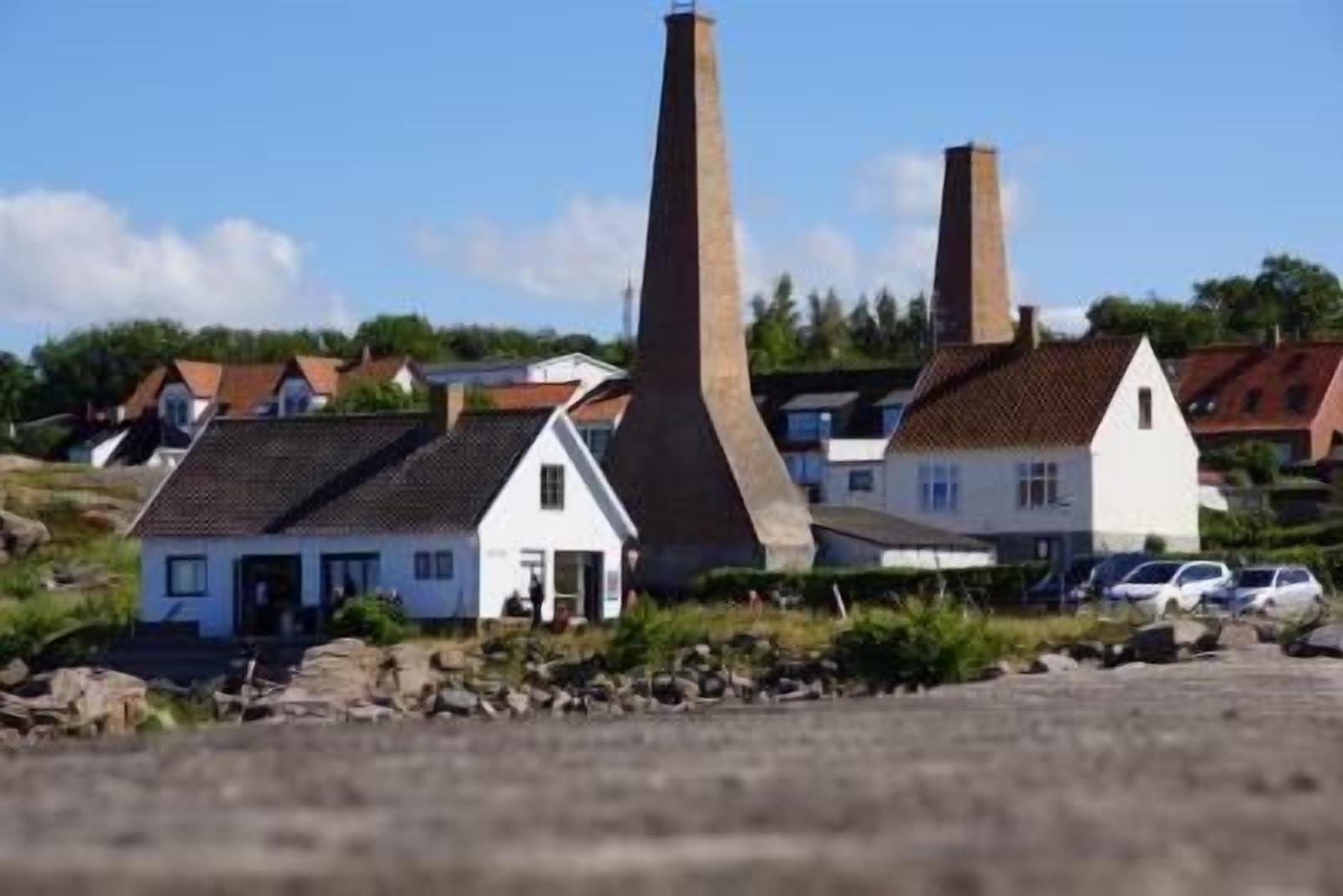 A coastal village with traditional houses, two prominent chimneys, and parked cars.
