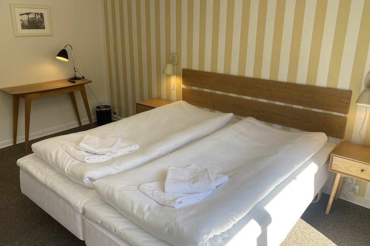 A neatly made bed with white linens and towels, a wooden headboard, a bedside table with a lamp, and a striped wall in the background.