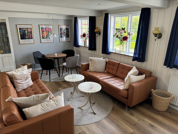 A cozy living room with a brown leather sofa, a round wooden coffee table, and a dining table with chairs. There are framed artworks on the wall, a vase with flowers on the windowsill, and a wicker basket on the floor.