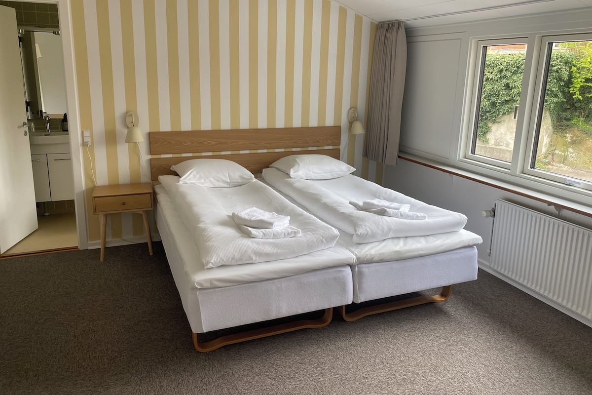 A double bed with white linens, a wooden headboard, a bedside table, a mirror, and a window with a view of greenery.