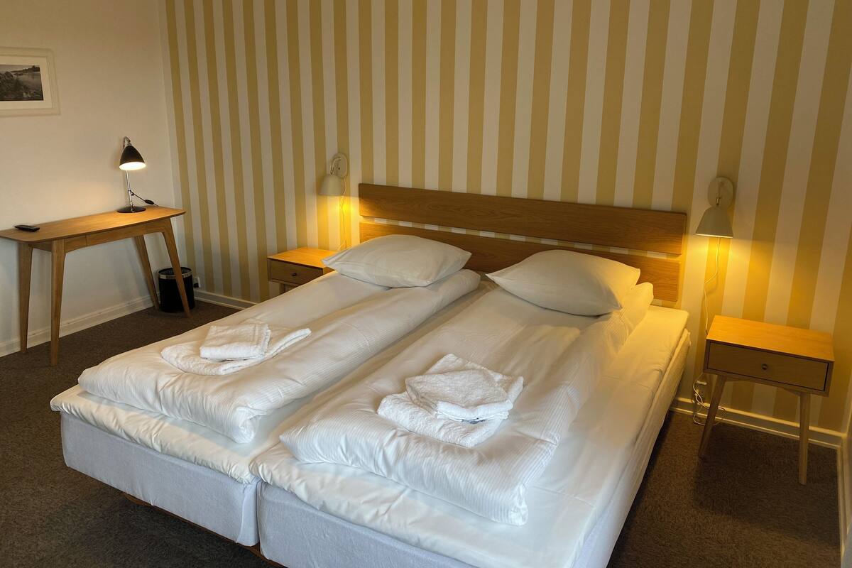 A double bed with white linens and towels, two bedside tables with lamps, a wooden desk, and a striped wall.