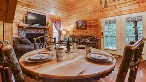 Ein Holzblockhaus-Innenraum mit einem runden Esstisch, Ledersesseln, einem Kamin, einem Fernseher und einer Tür mit Blick ins Freie.